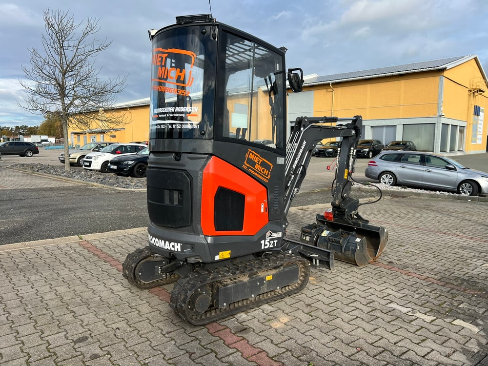 Schwarzer und oranger Minibagger auf einem Parkplatz vor Gebäuden und Autos.