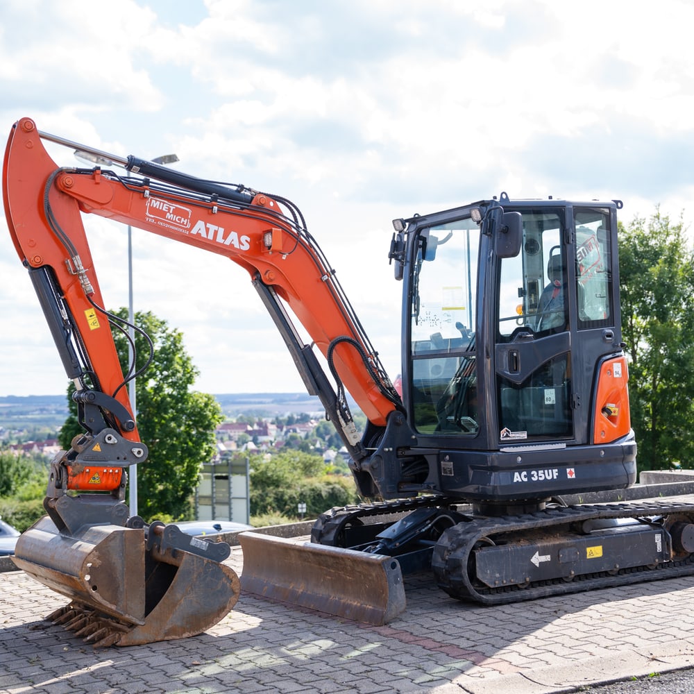 Orangefarbener Atlas Minibagger AC 35UF auf einem gepflasterten Parkplatz mit Bäumen und Häusern im Hintergrund.