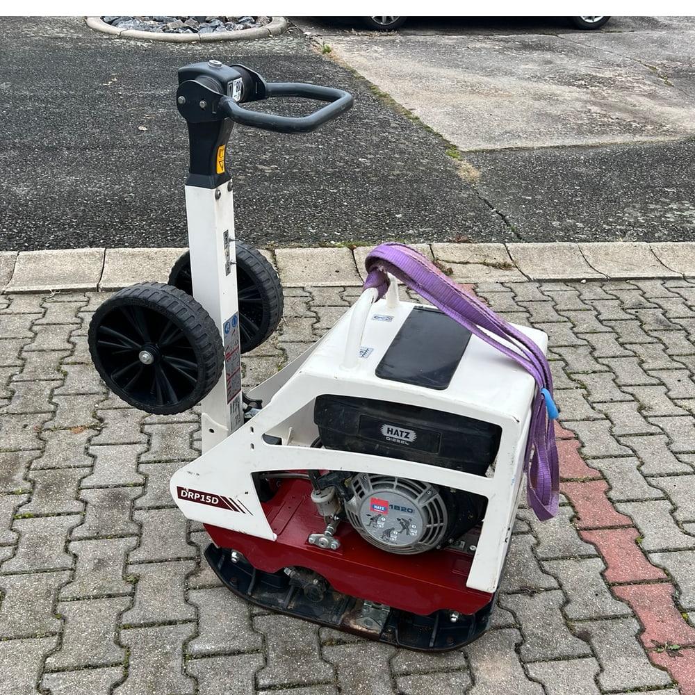 Weiße und rote Rüttelplatte mit Griff, Rädern und lila Spanngurt, auf Pflastersteinen.