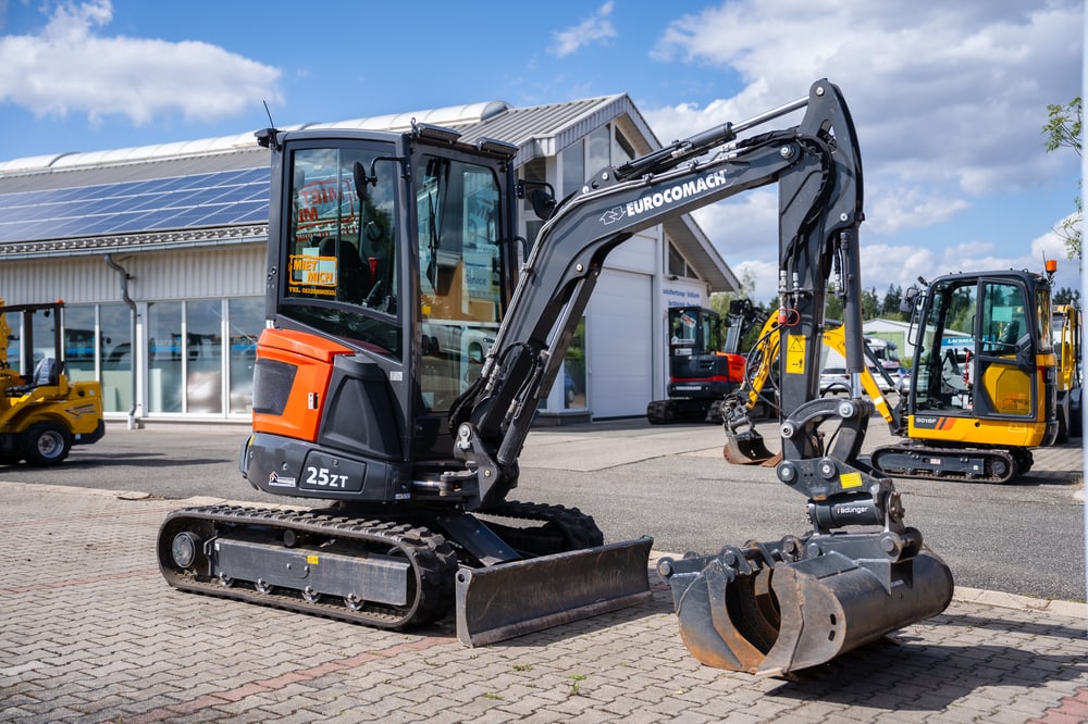 Schwarzer Eurocomach 25ZT Minibagger mit Greiferschaufel, auf einer Baustelle mit weiteren Maschinen und Gebäude.