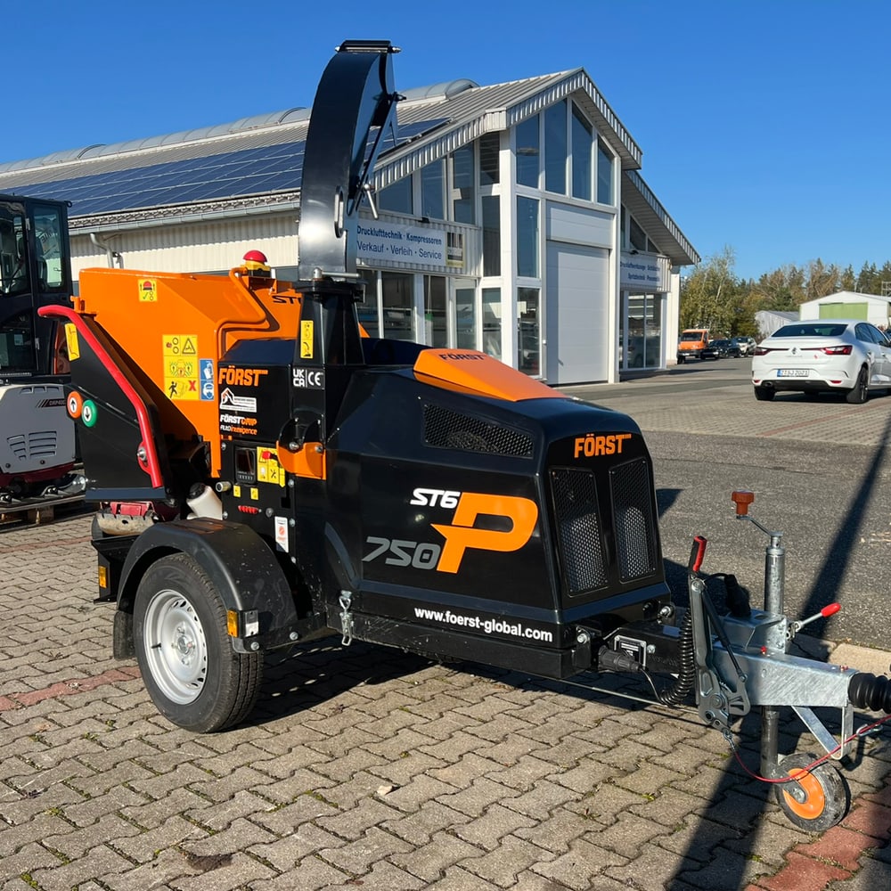 Schwarzer und oranger Först ST6 750 Holzhacker auf Anhänger, vor einem Gebäude und einem weißen Auto.