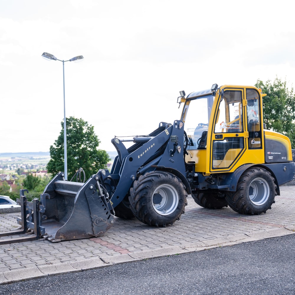 Gelber und grauer Radlader mit Gabeln und Schaufel, geparkt auf einem Pflastersteinweg.